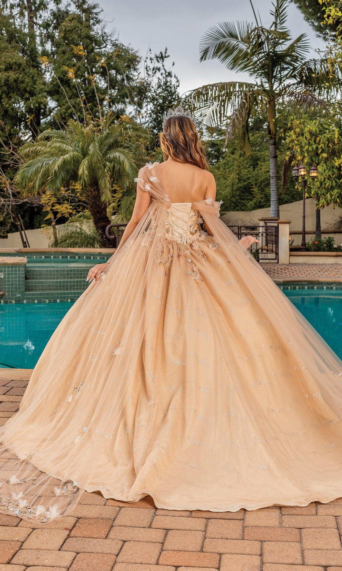 A woman in the Quinceanera Dress 1844 By Dancing Queen, featuring floral details and a sweetheart neckline, stands by a pool in an outdoor garden setting, facing away from the camera.